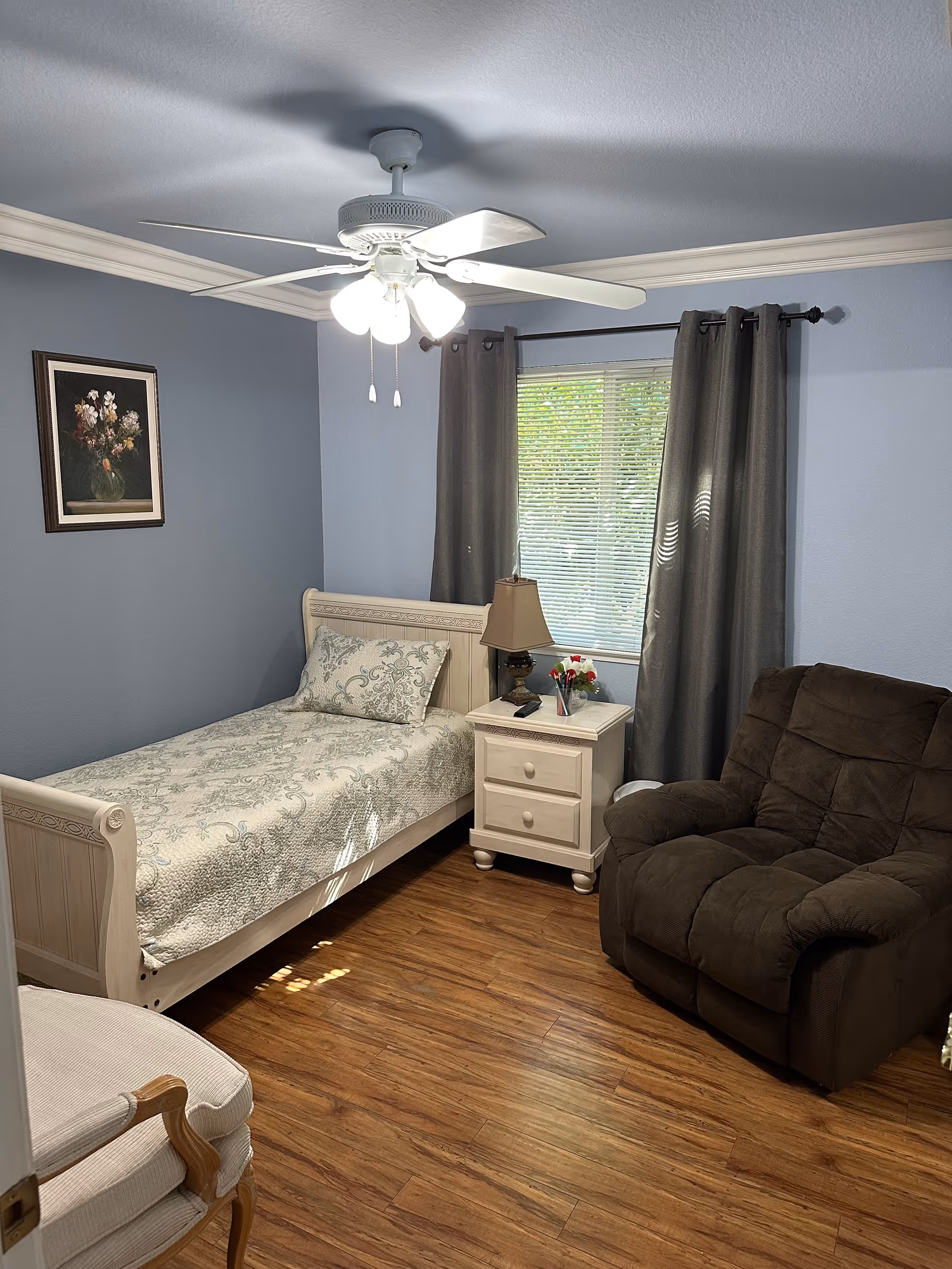 Light-blue bedroom with a single bed, nightstand and lamp, brown recliner, ceiling fan, and a window with gray curtains.