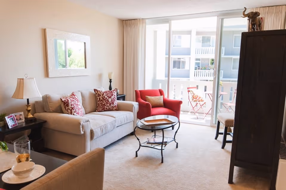 Bright living room with a beige sofa, red armchair, glass-top coffee table and sliding glass doors opening to a balcony.