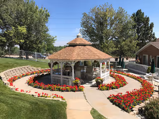 A gazebo with a shingled roof surrounded by a circular flower bed filled with red flowers. The gazebo is located in a well-maintained outdoor area with green grass, trees, and a paved walkway leading to it. There is a brick building visible on the right side and a metal fence in the background.