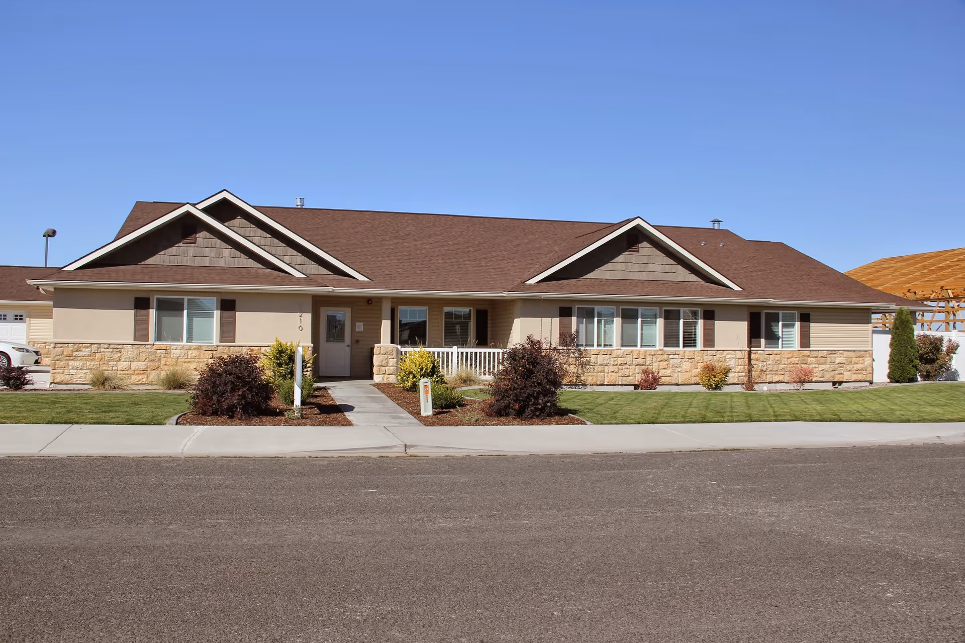 Single-story building with a brown roof, beige walls, and stone accents. The building has multiple windows with brown shutters and a small porch with a white railing. There is a well-maintained lawn and landscaping with bushes in front of the building under a clear blue sky.