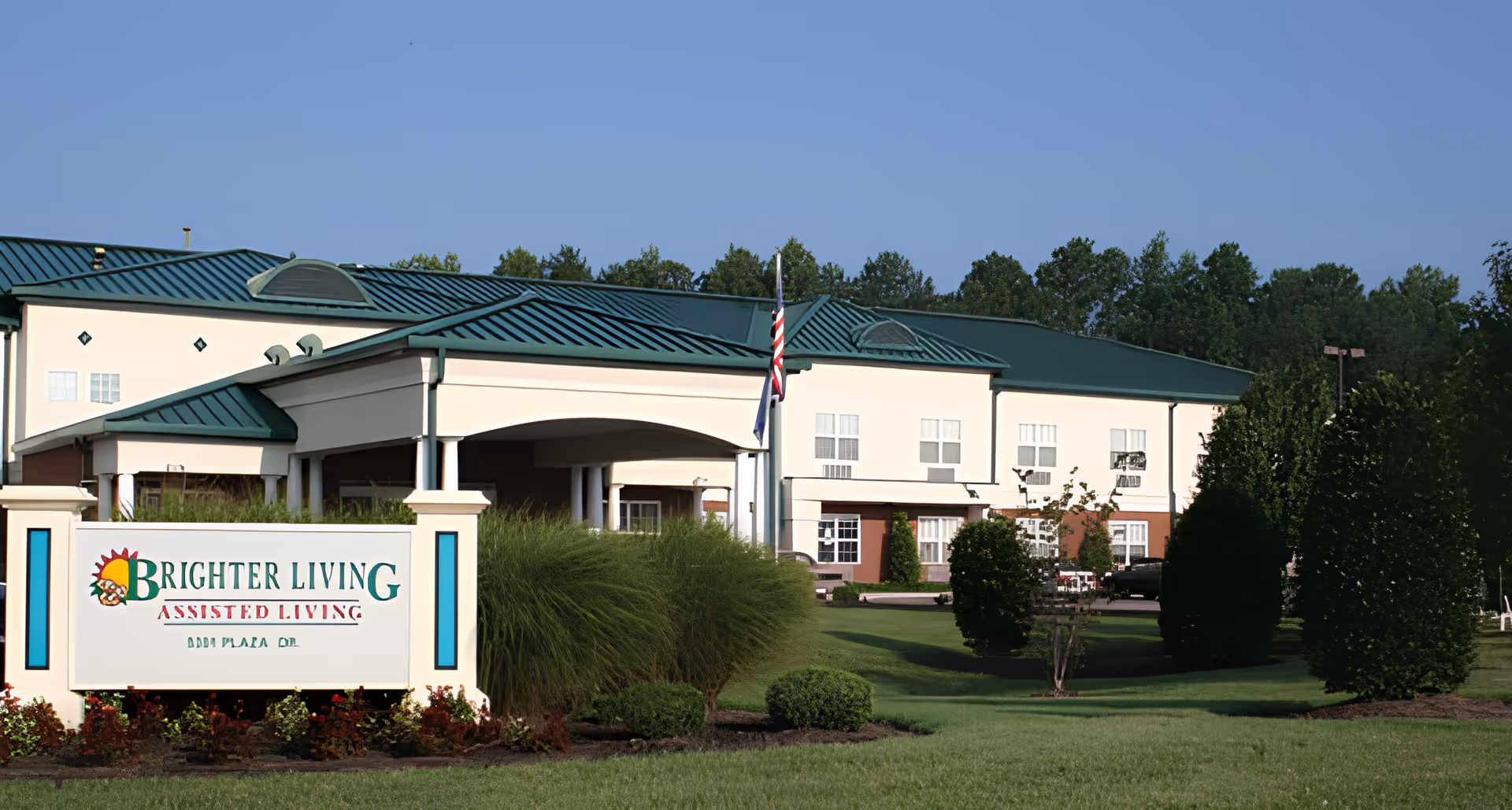 Exterior view of Brighter Living Assisted Living facility with a green roof, beige walls, and a well-maintained lawn with bushes and trees. A sign with the facility's name is visible in the foreground, and an American flag is flying near the entrance.
