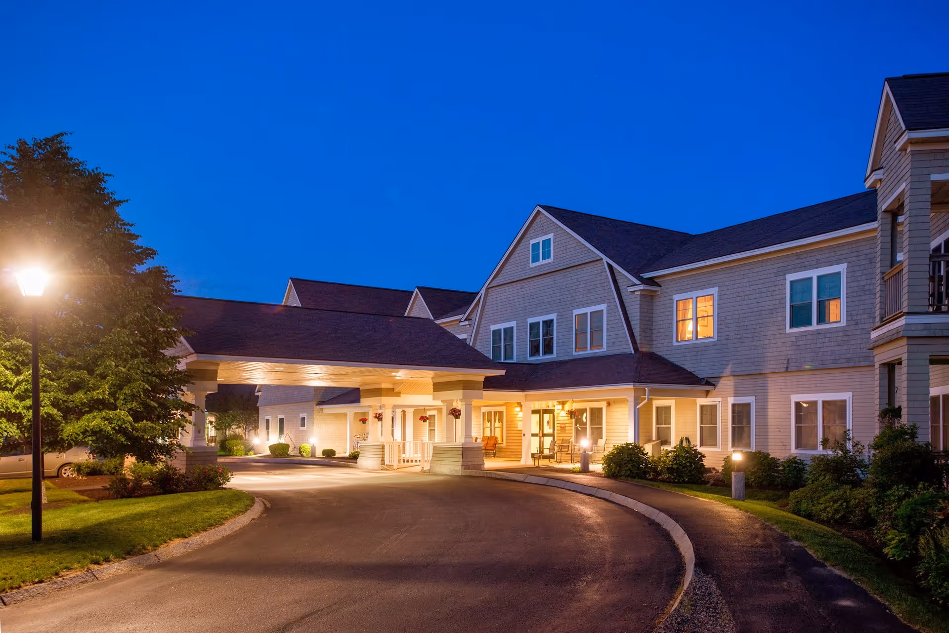 Lit entrance and porte-cochère of a multi-story senior living building at dusk with a curved driveway.