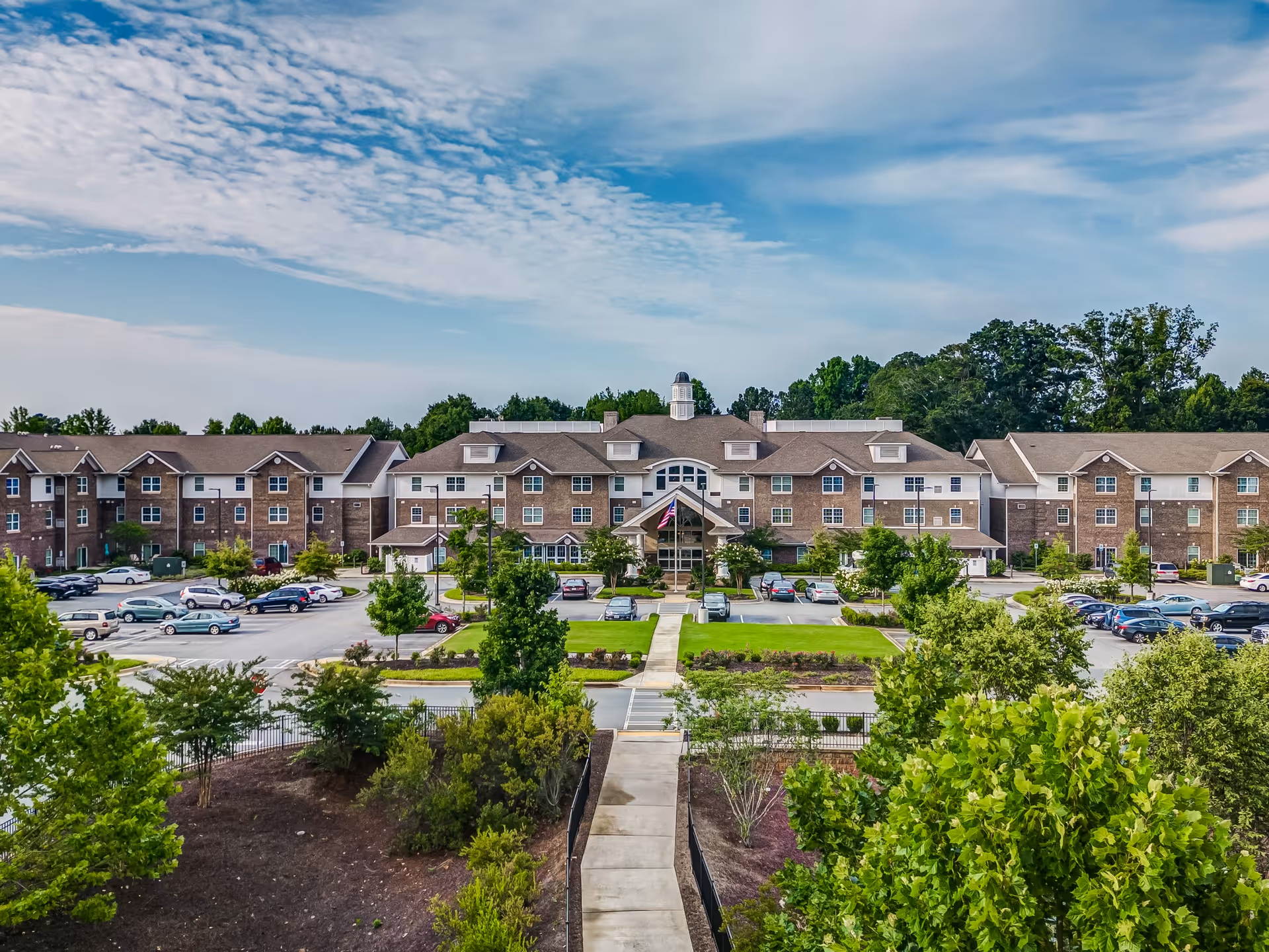 Front exterior of a large three-story brick retirement facility with a central entrance, parking lot, and landscaped grounds with a walkway leading to the building.