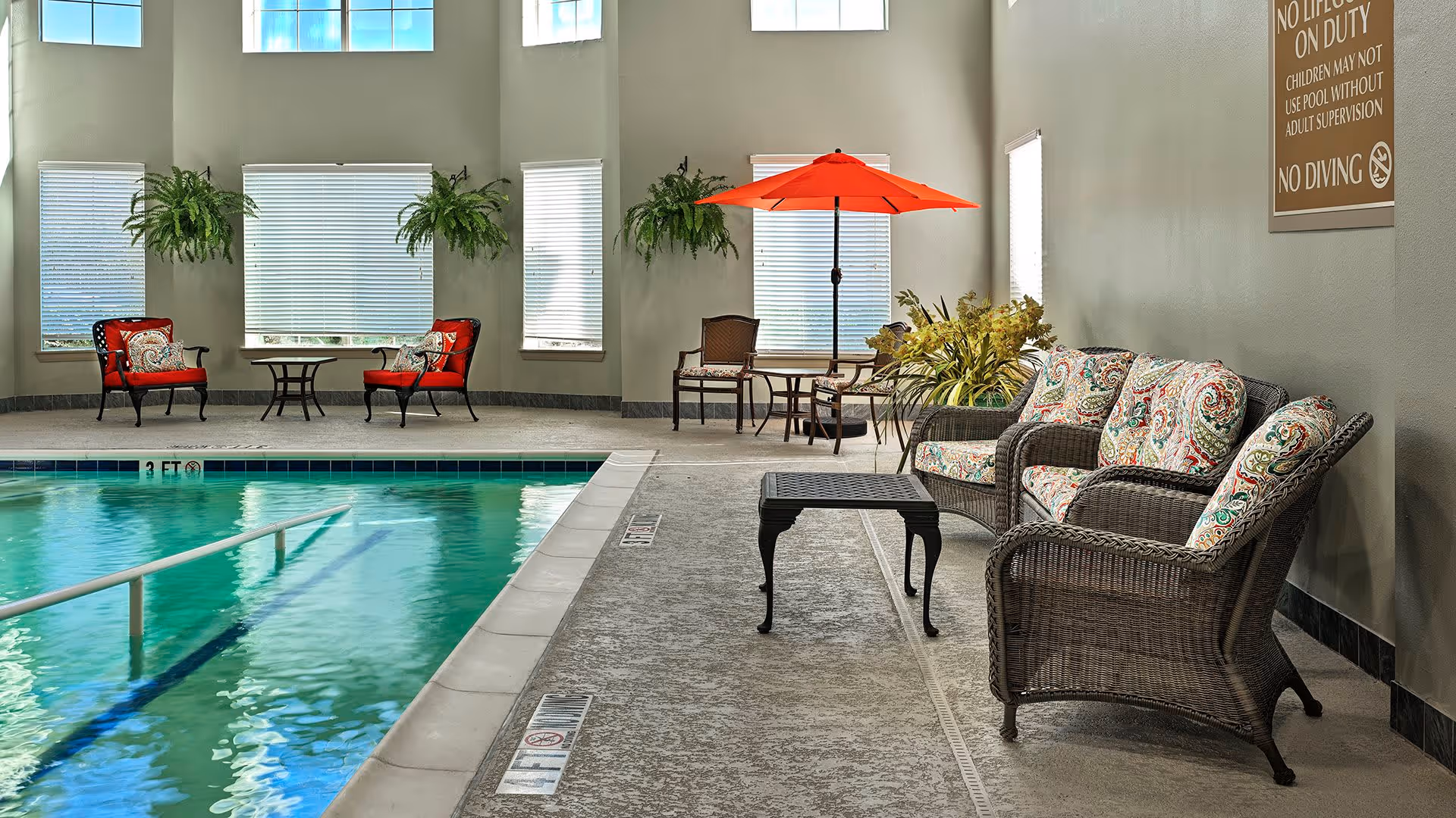 Indoor swimming pool area with clear water, surrounded by seating including wicker chairs with patterned cushions, a small table, and red cushioned chairs near windows. There is a red umbrella over a table with chairs, potted plants, and a sign on the wall with pool rules.