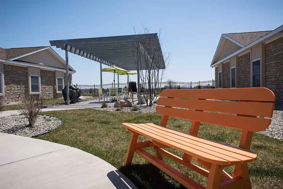 Orange bench in a courtyard with a pergola, patio seating and stone-clad buildings under a clear blue sky.