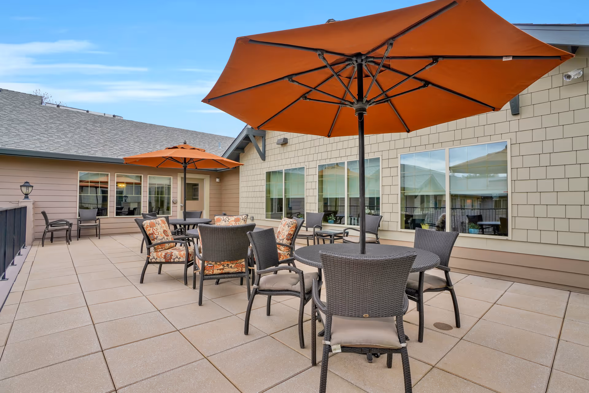 Outdoor patio area with multiple round tables and chairs, some with floral cushions, shaded by large orange umbrellas. The patio is adjacent to a building with beige siding and several windows reflecting the outdoor scene.