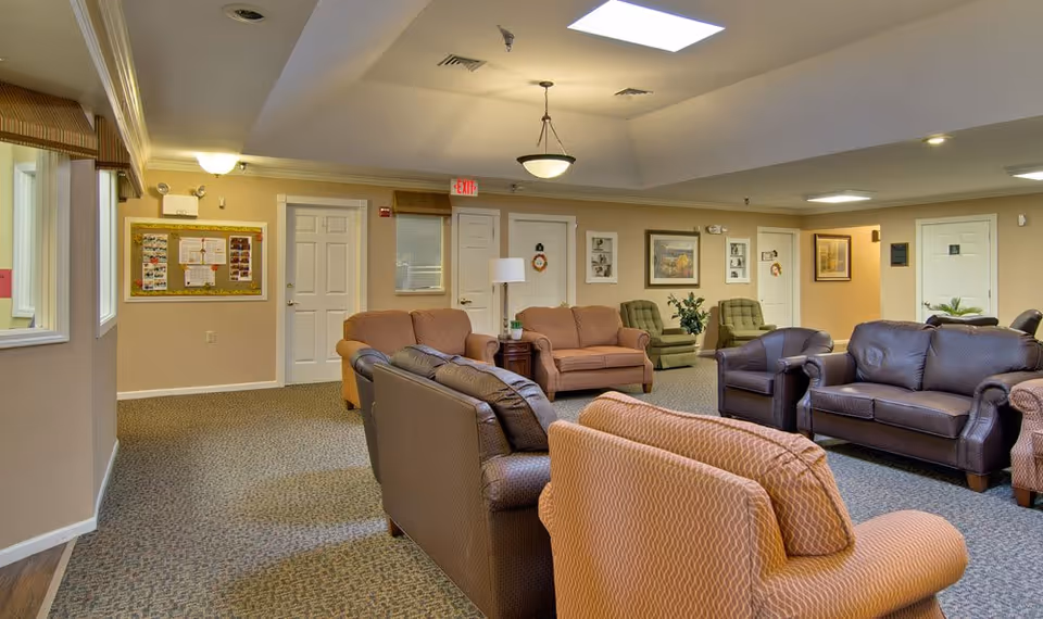 A spacious senior living facility common area with multiple sofas and armchairs arranged for socializing. The room has beige walls, carpeted floors, framed artwork on the walls, and ceiling lights including a hanging light fixture. Several doors and a bulletin board are visible along the walls.