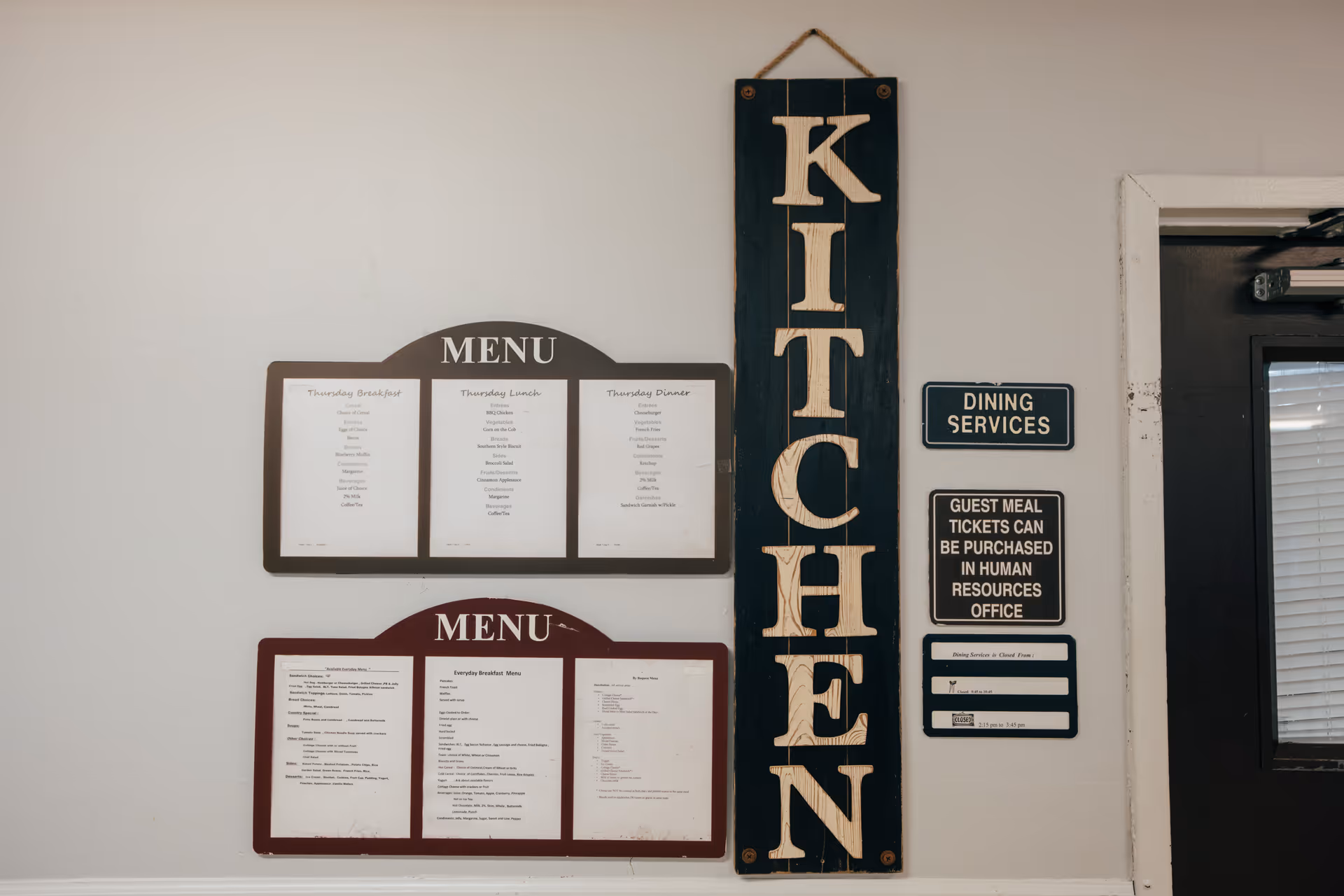 Wall with two menu boards displaying meal options, a vertical wooden sign with the word 'KITCHEN', and several informational signs about dining services and guest meal tickets near a door.