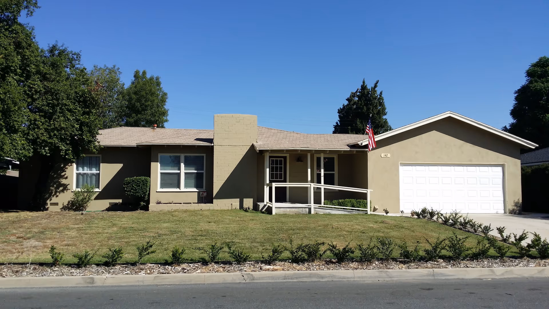 Single-story residential building with a beige exterior, a white garage door, an American flag near the entrance, and a wheelchair ramp leading to the front door. The house is surrounded by a lawn with some bushes and trees under a clear blue sky.