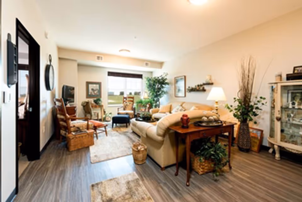 A cozy living room with a beige sectional sofa, wooden chairs, a small wooden table with decorative items, a floor lamp, and various plants. The room has wood flooring, a large window with blinds, and framed artwork on the walls.