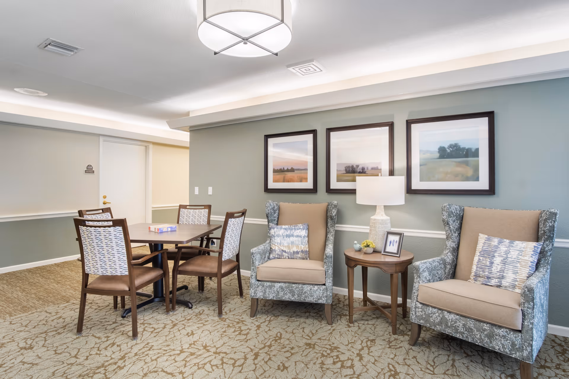 A cozy sitting area in a senior living facility with two patterned armchairs featuring beige cushions and decorative pillows, a small wooden side table with a lamp, framed landscape artwork on the wall, and a table with four chairs in the background.