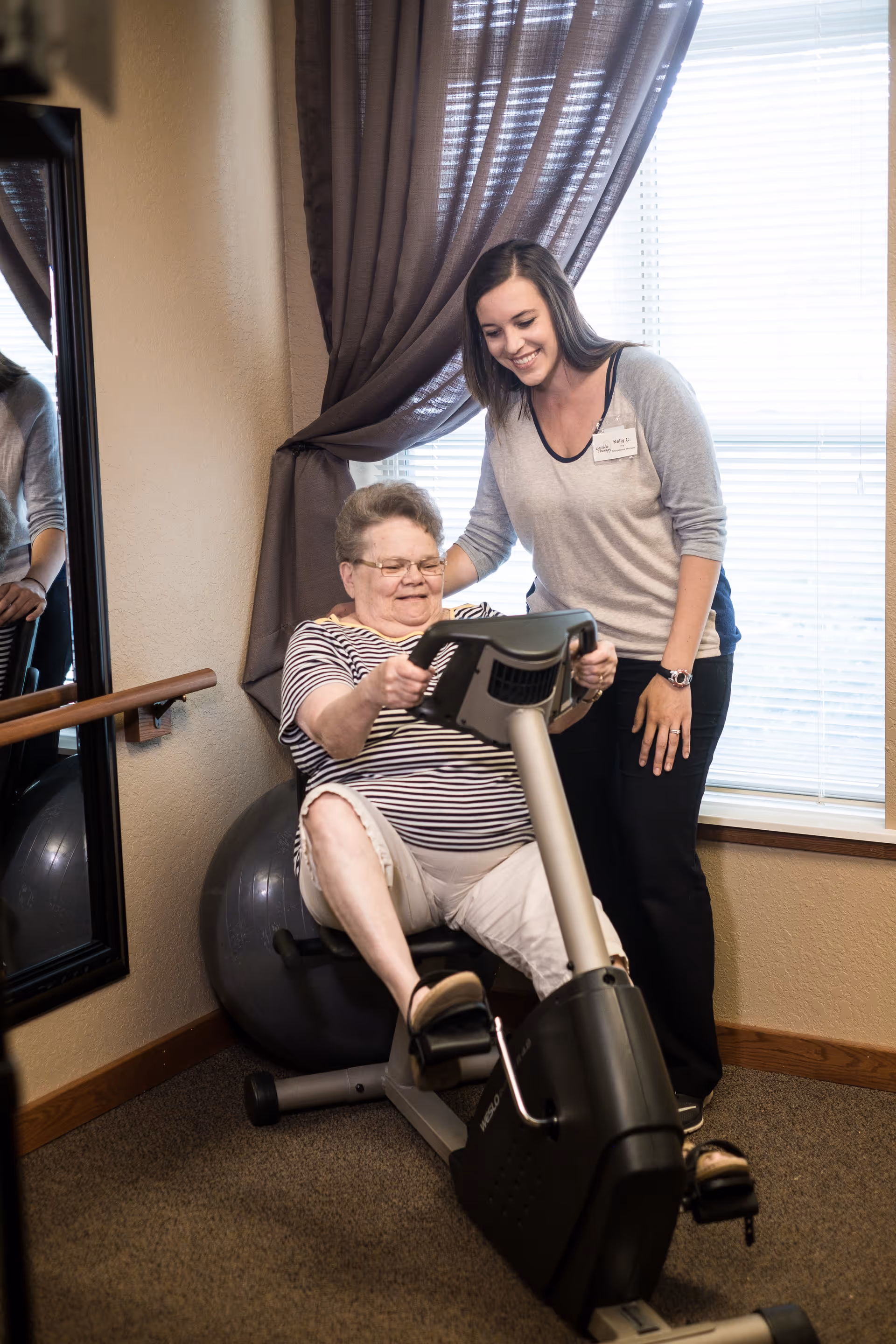 An elderly woman is seated on an exercise bike, smiling and pedaling while a younger woman stands beside her, offering support and encouragement in a room with a large window and brown curtains.