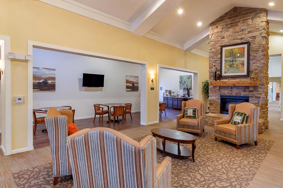 A cozy assisted living facility common area featuring a stone fireplace with a framed painting above it. The room has comfortable armchairs with striped upholstery and a wooden coffee table on a patterned carpet. Adjacent rooms have tables and chairs, a wall-mounted TV, and a refreshment station with coffee and snacks. The walls are painted in warm tones with soft lighting.