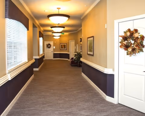 A well-lit hallway in a senior living facility with beige and dark purple walls, carpeted floor, ceiling lights, framed pictures on the walls, and doors decorated with autumn wreaths.