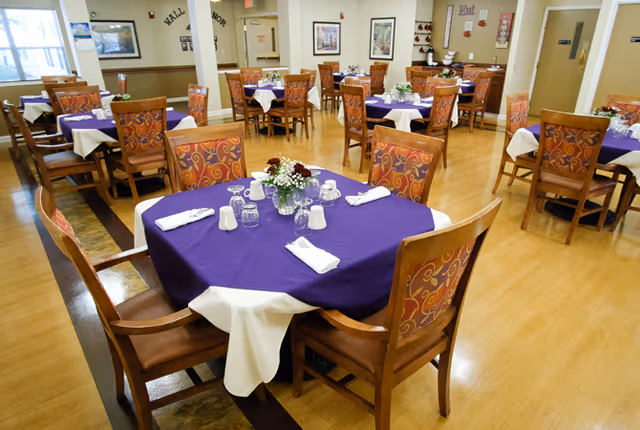 A dining room with multiple wooden tables covered with purple and white tablecloths. Each table is set with white napkins, glasses, salt and pepper shakers, and floral centerpieces. The room has wooden flooring and walls decorated with framed pictures and small shelves. There are several wooden chairs with patterned upholstery around the tables.