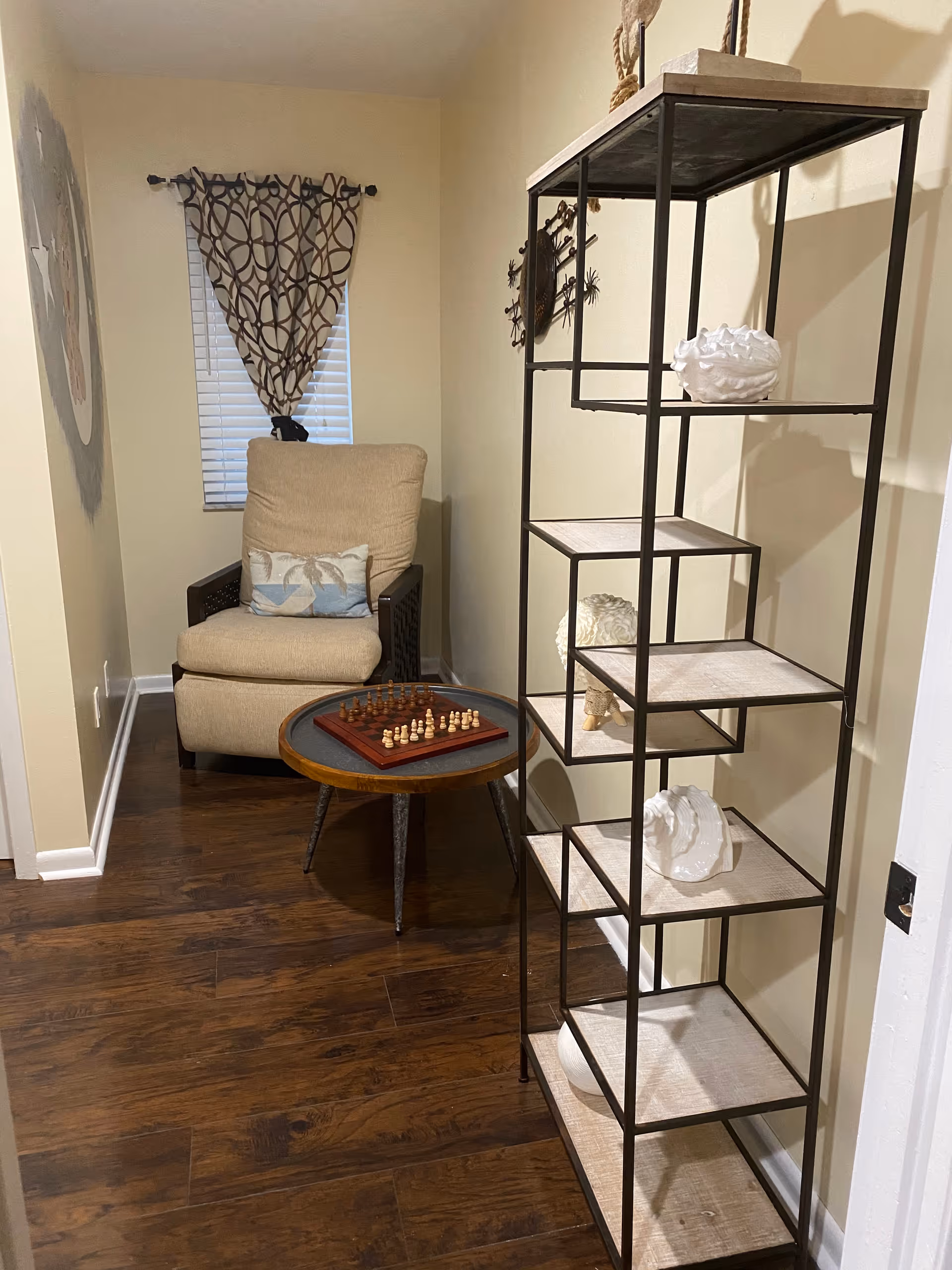 Small sitting area with an armchair, a round side table holding a chessboard, and a tall open shelving unit against a wall.