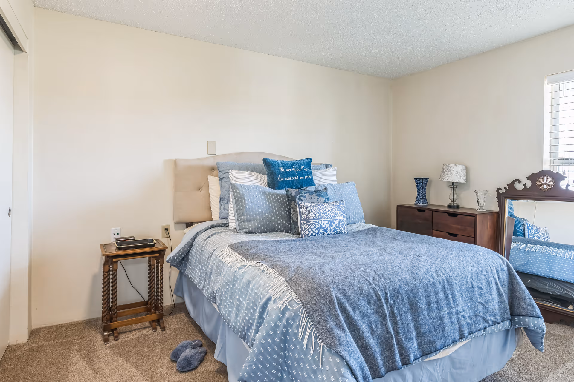 A cozy bedroom with a neatly made bed featuring multiple blue and white pillows and a blue blanket. There is a small wooden side table with a phone and charger on the left side of the bed. On the right side, there is a wooden dresser with a blue vase, a lamp, and a glass decoration. A large ornate mirror leans against the wall near a window with blinds, and a pair of blue slippers is on the carpeted floor.