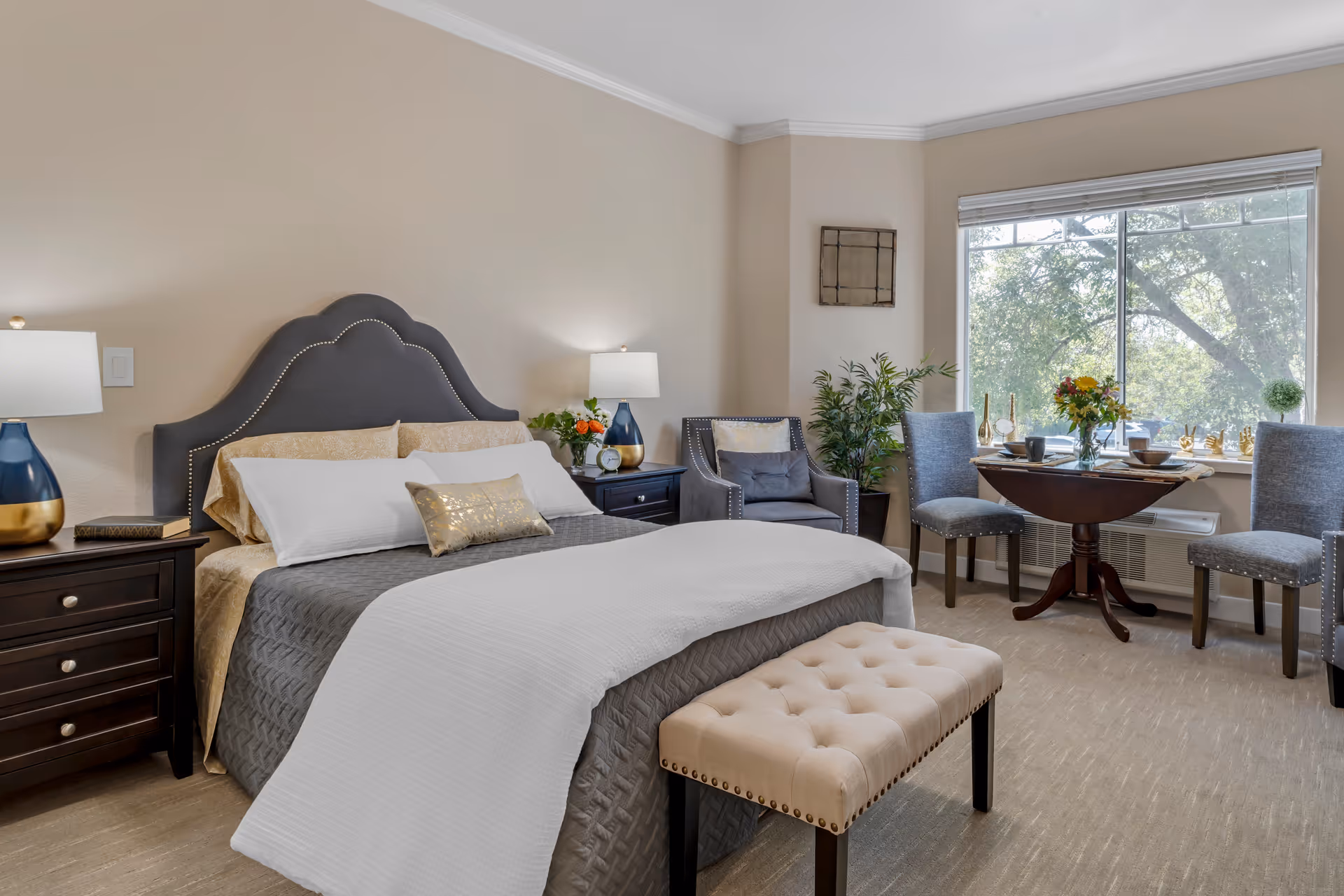 Well-lit furnished bedroom with a tufted bench, queen bed, nightstands, seating area and a small dining table by a large window.