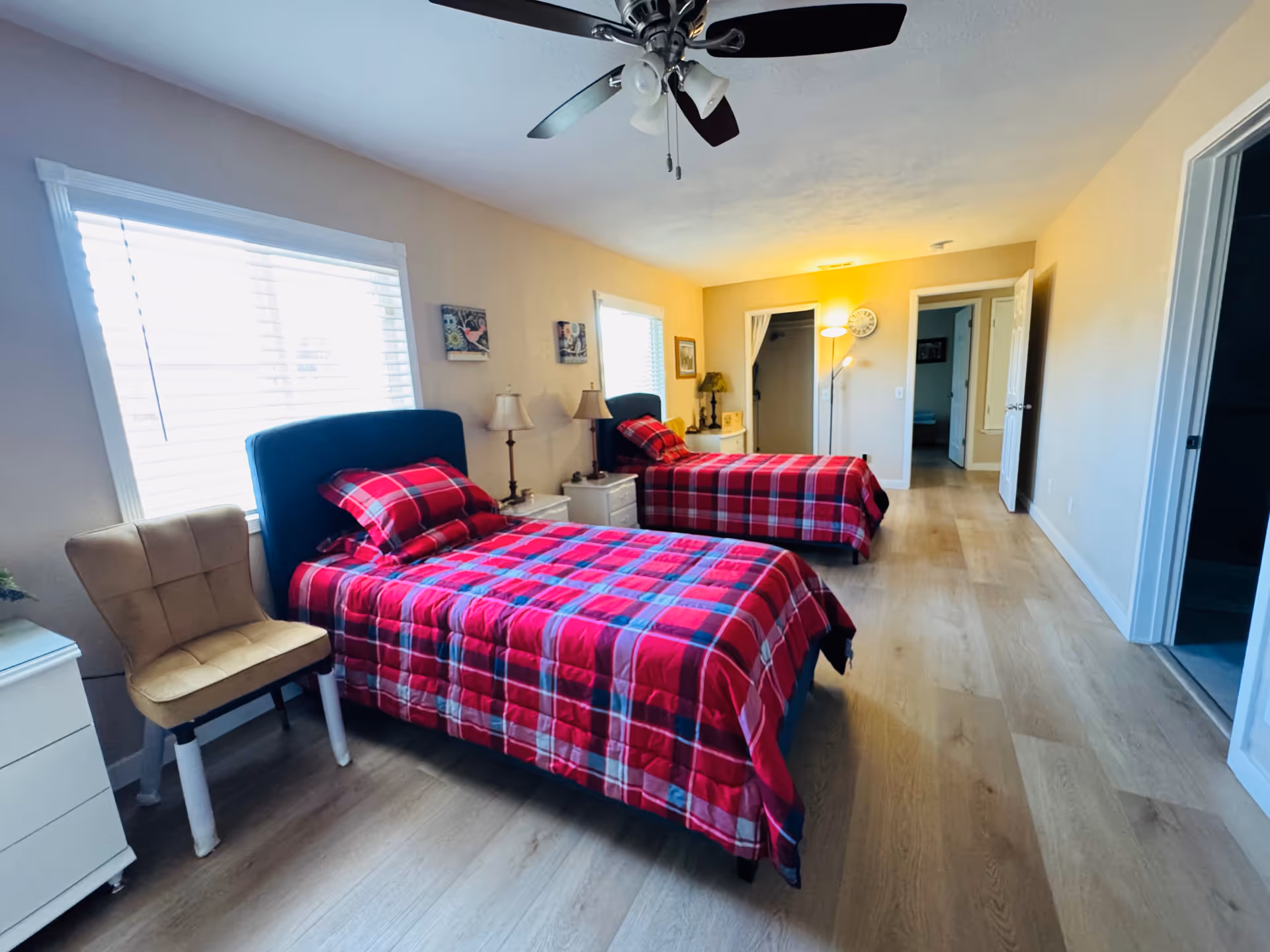 A bright bedroom with two single beds covered in red and black plaid bedding. Each bed has a matching pillow and a blue upholstered headboard. There are two windows with white blinds letting in natural light. Between the beds is a white nightstand with two lamps. A beige upholstered chair is placed near the window. The room has light wood flooring, a ceiling fan with lights, and a floor lamp near the far wall. Doors lead to other rooms or closets.