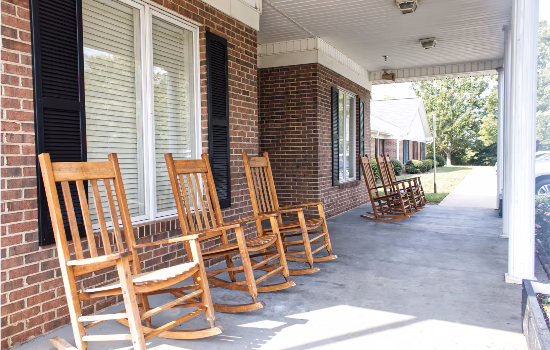 A covered outdoor porch area with six wooden rocking chairs lined up against a brick wall with windows. The porch has a concrete floor and white columns supporting the roof. There is greenery and a sidewalk visible in the background.