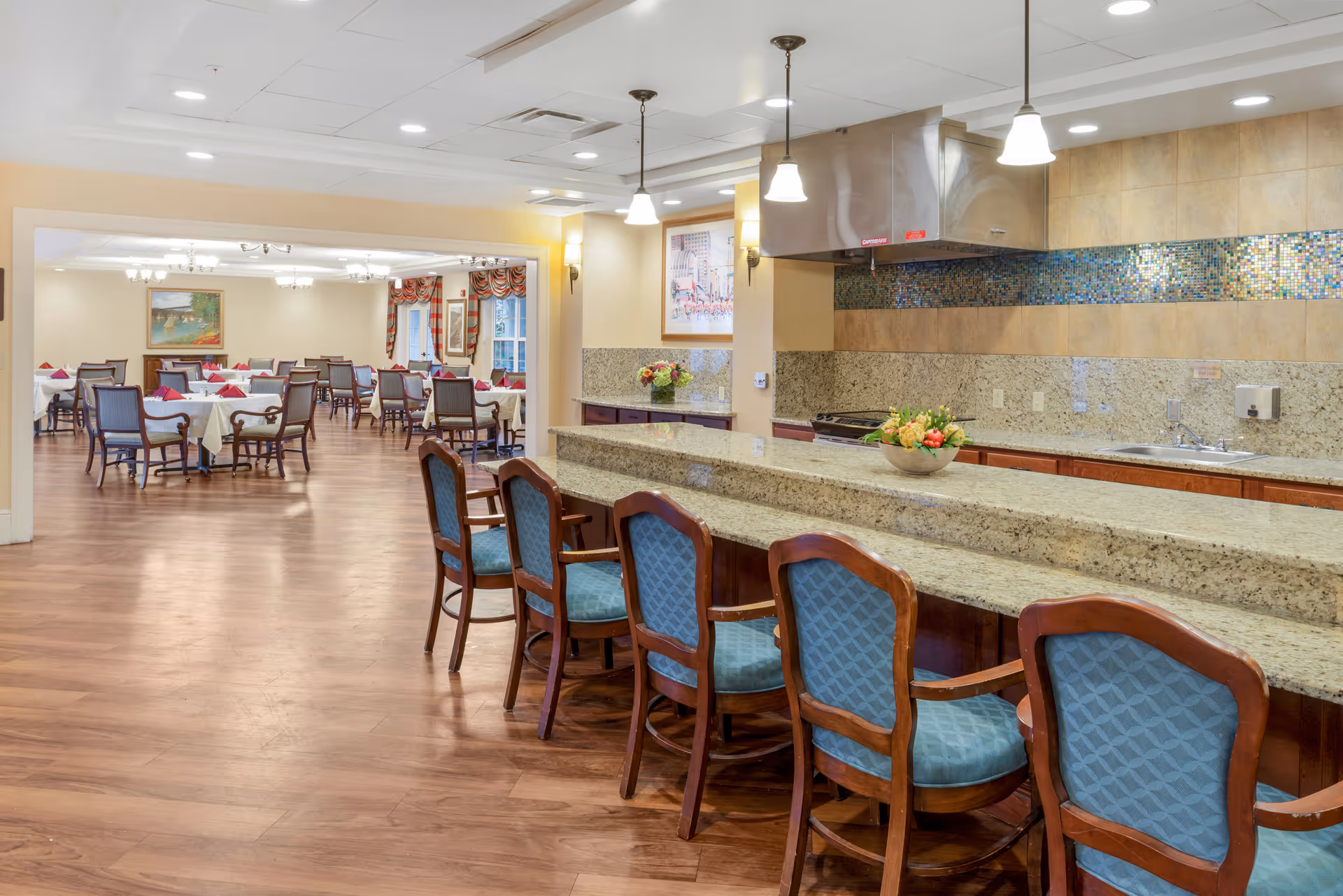 A spacious dining area in a senior living facility featuring a long granite countertop with six wooden chairs upholstered in blue fabric. Behind the counter is a kitchen area with a stainless steel range hood, mosaic tile backsplash, and a sink. In the background, there is a large dining room with multiple tables covered with white tablecloths and set with red napkins, surrounded by wooden chairs. The room has wooden flooring, warm lighting, and framed artwork on the walls.