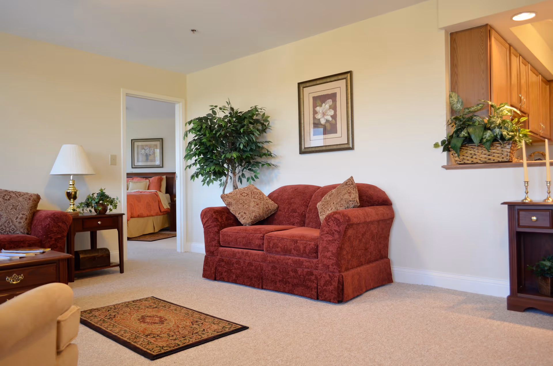 A cozy living room with a red patterned loveseat and matching armchair, a small wooden side table with a lamp and plant, a framed flower picture on the wall, a potted plant in the corner, and a view into a bedroom with a bed covered in red and beige bedding.