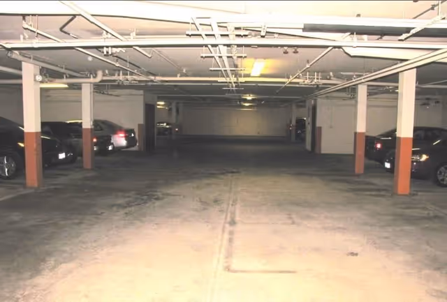Indoor parking garage with several parked cars along the sides and concrete pillars supporting the ceiling with exposed pipes and lighting fixtures.