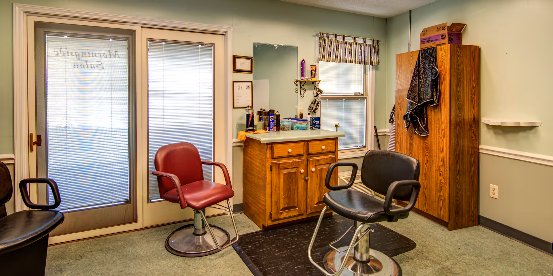 Interior of a small salon area with two salon chairs, one red and one black, positioned in front of a wooden cabinet with hair care products on top. There is a large mirror above the cabinet, a window with blinds and a valance, and a wooden storage cabinet with towels hanging on the side. The room has light green walls with white trim and a door with frosted glass panels.