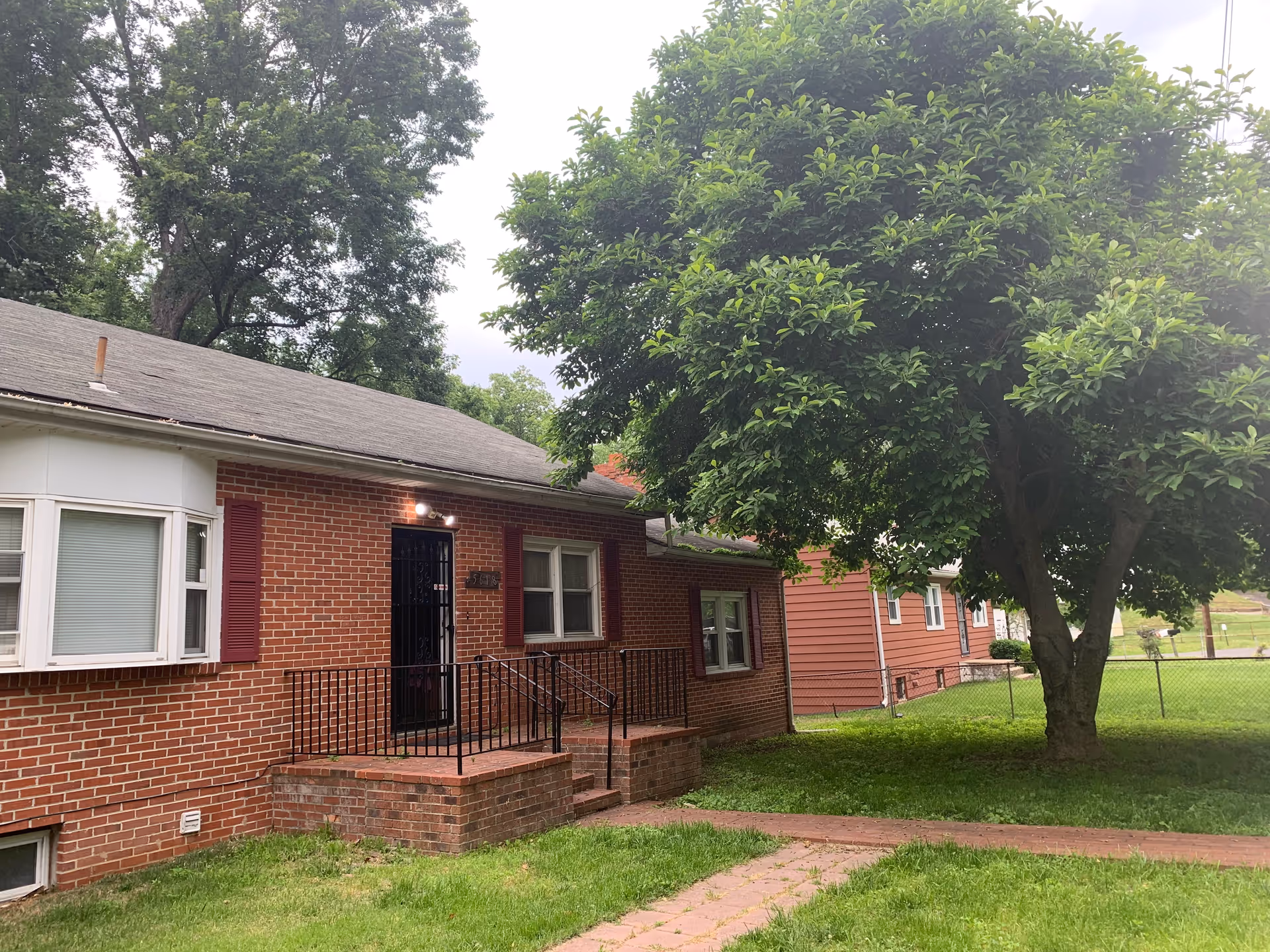 Exterior view of a single-story brick house with a small front porch and black metal railing. There is a large leafy tree on the right side and a green lawn with a paved walkway leading to the porch. The house has white-framed windows with red shutters and a gray shingled roof. Another similar house is visible in the background.