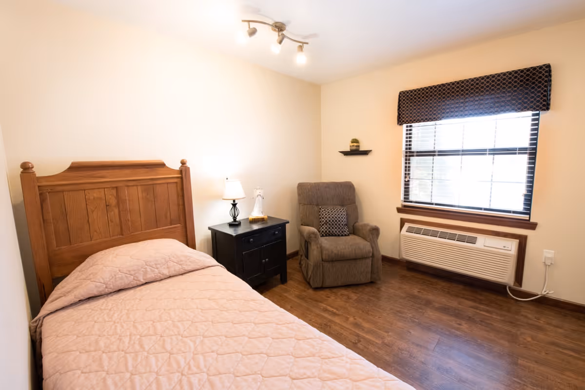 A simple bedroom with a single wooden bed, nightstand and lamp, a recliner chair, and a window with an air conditioner.