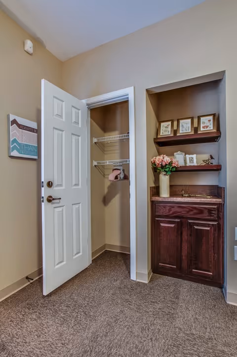 Interior corner of a room with an open white door revealing a small closet with wire shelving. Next to the closet is a built-in wooden cabinet with a countertop and two shelves above it, decorated with framed pictures and a vase of pink flowers. The walls are beige and the floor is carpeted.
