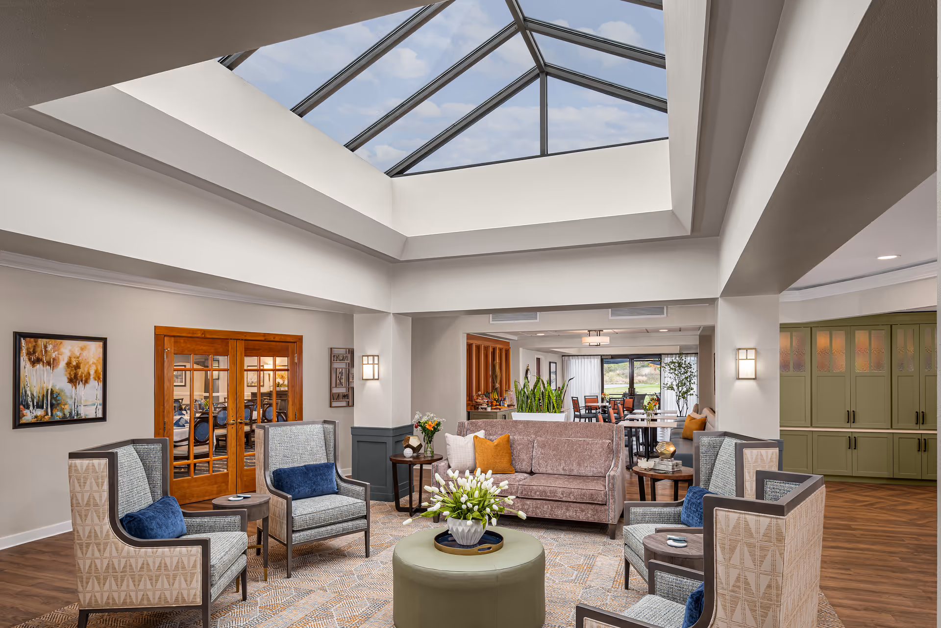A bright and spacious senior living facility common area with a large skylight ceiling. The room features a comfortable seating arrangement including a sofa with orange and white pillows, four patterned armchairs with blue cushions, a round green ottoman with a vase of white flowers, and side tables. In the background, there is a dining area with tables and chairs, green cabinetry, and large windows letting in natural light. The walls are decorated with framed artwork and the flooring is wood with a patterned rug.