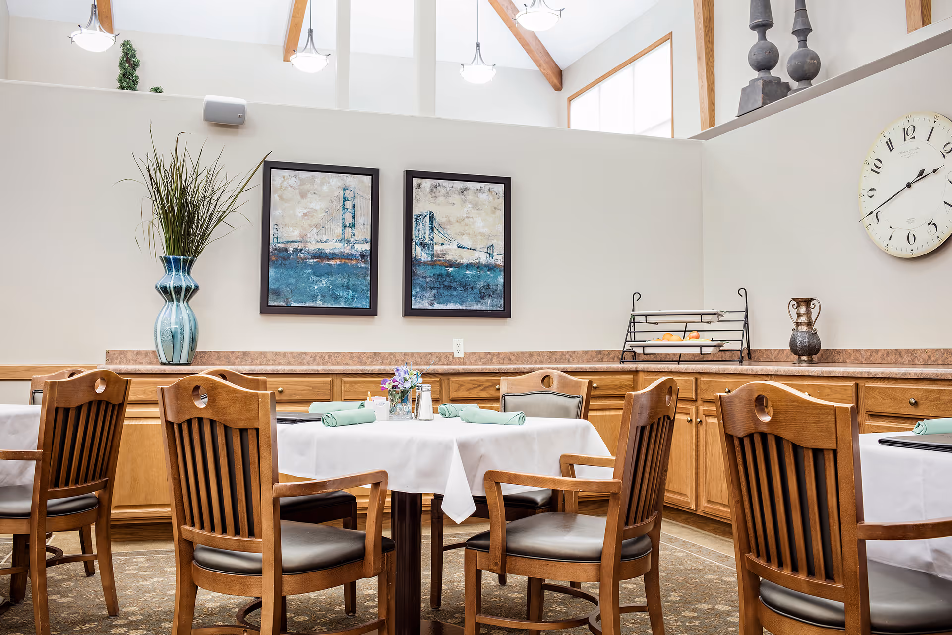 Bright dining room with wooden chairs and tables set with white tablecloths, artwork on the wall, and a large clock.