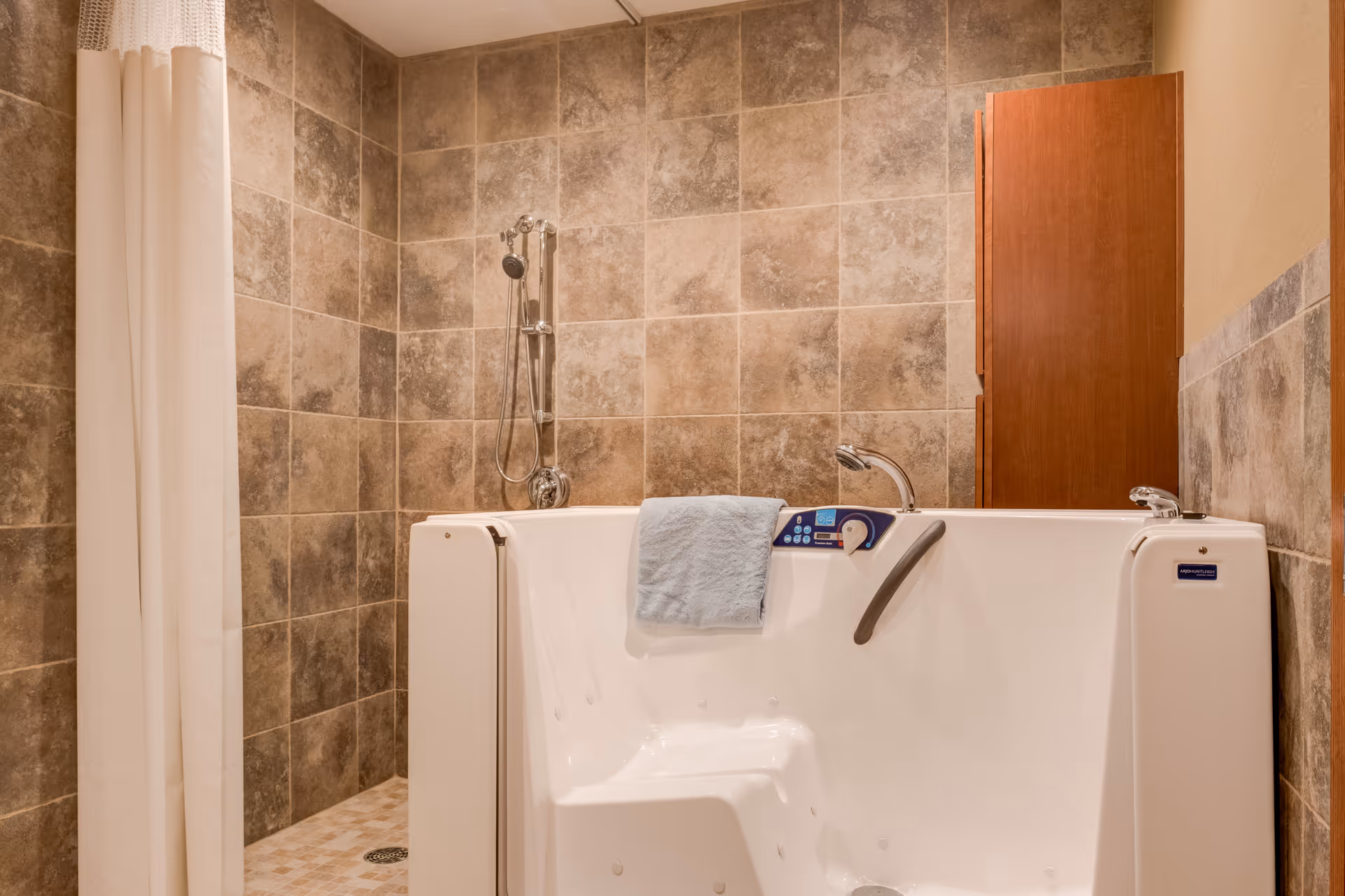 A tiled bathroom featuring a walk-in bathtub with a handheld shower, towel draped over the tub, and a wooden storage cabinet.