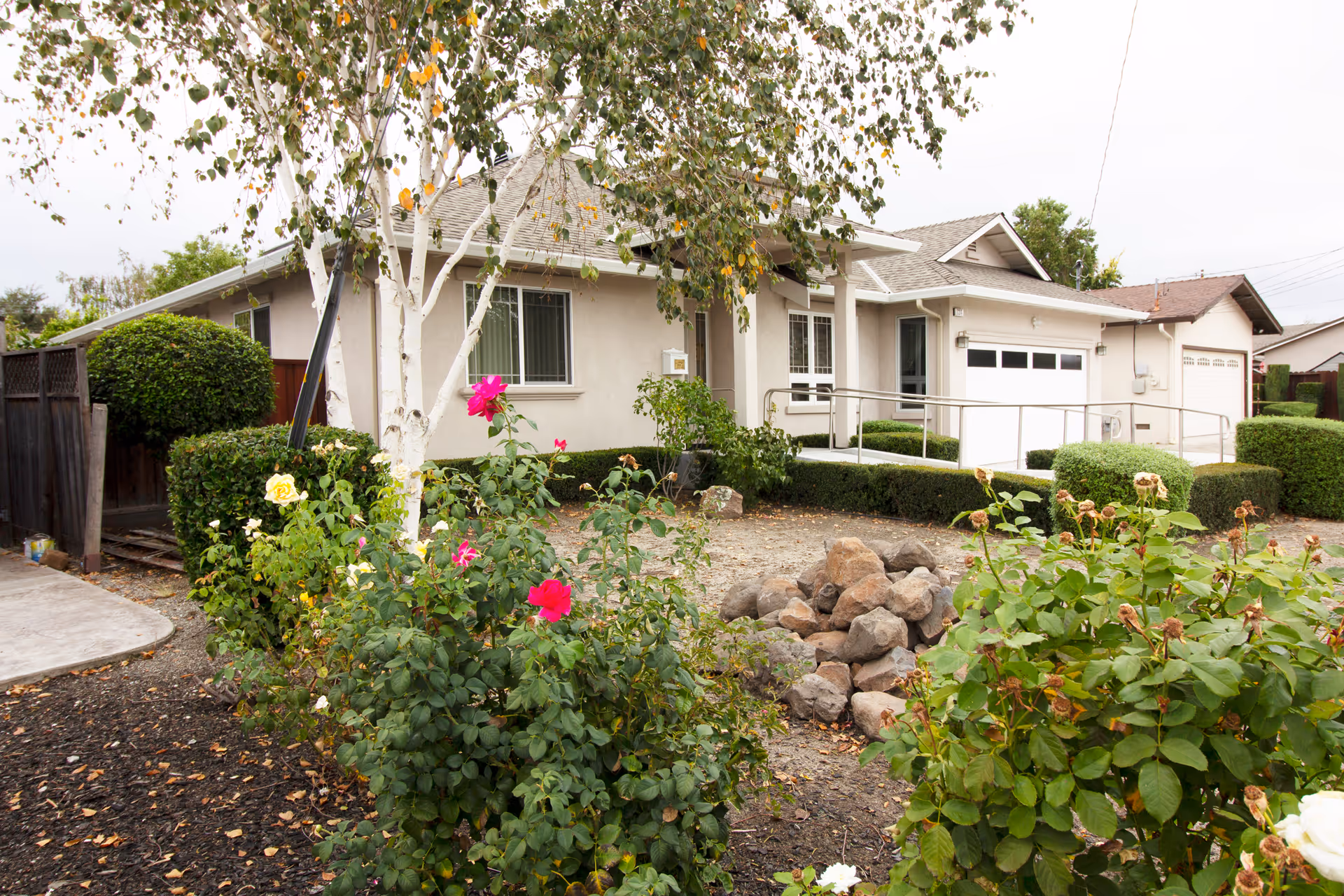 Exterior view of a single-story residential building with beige walls and a gray roof, surrounded by green bushes and flowering plants. A concrete pathway leads to the entrance, which has a metal handrail ramp. There is a garage door on the right side of the building. The sky is overcast.