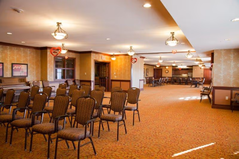 A large, carpeted room with rows of upholstered chairs arranged facing forward. The room has warm lighting with ceiling fixtures and decorative wallpaper. There are red heart-shaped decorations hanging from the ceiling. In the background, there is a piano against the wall and additional seating areas further back.