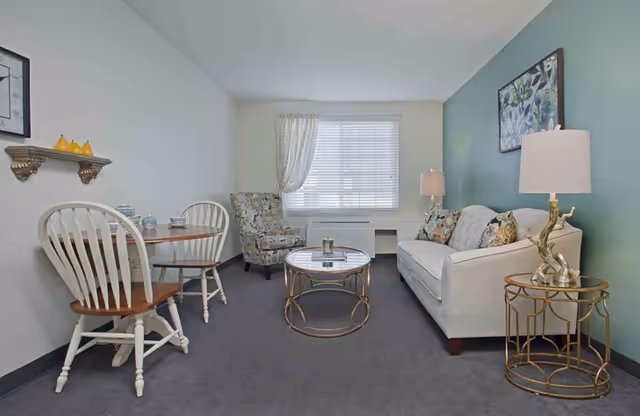 A cozy living room with a white sofa adorned with patterned cushions, a round glass coffee table with a gold frame, and a matching side table with a lamp. There is a floral armchair near a window with white blinds and a sheer curtain. A small wooden dining table with two white chairs is positioned against the opposite wall, which has a small shelf with decorative items and a framed picture above it. The walls are painted in light colors with one accent wall in green.