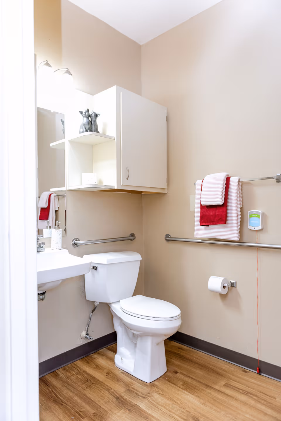 A clean and accessible bathroom with a white toilet, a wall-mounted sink, and a white cabinet above the toilet. There are grab bars on the walls near the toilet and sink. A towel rack holds a red and white towel, and a soap dispenser is mounted on the wall. The floor has a wood-like finish.
