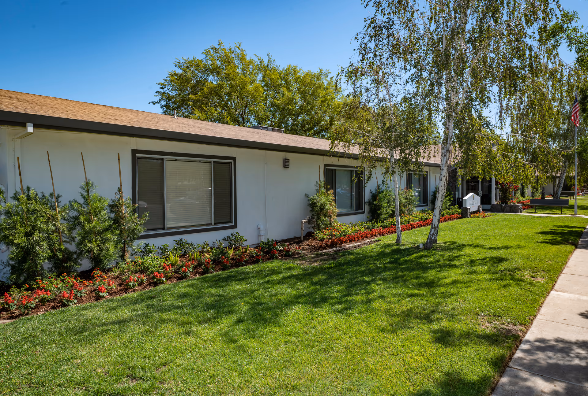 Exterior view of a single-story building with white walls and a brown roof, surrounded by a well-maintained lawn, flower beds with red flowers, small bushes, and several trees. A sidewalk runs along the right side of the image, and an American flag is visible in the background near the entrance.