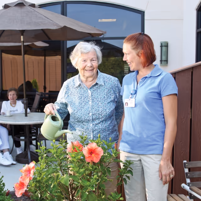 An elderly woman watering flowering plants with a green watering can while smiling and standing next to a female caregiver in a blue polo shirt. They are outside on a patio area with a large umbrella and another elderly woman sitting at a table in the background.