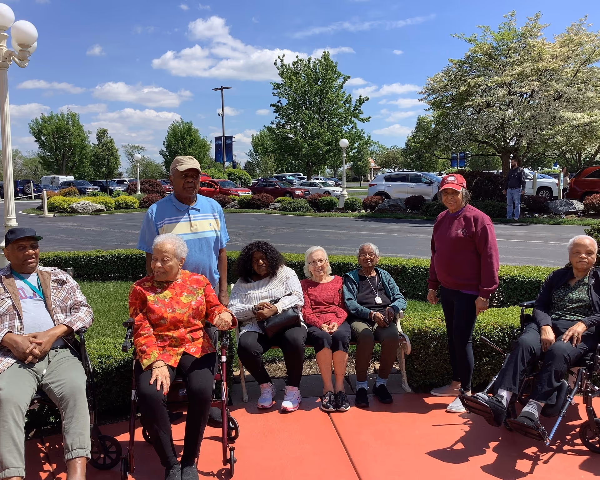 A group of elderly people sitting and standing outside on a sunny day near a parking lot with trees and bushes in the background. Some are seated on benches and others in wheelchairs, enjoying the outdoor setting.