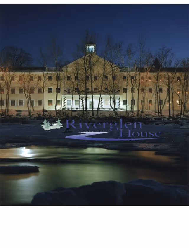 Night view of a large, multi-story building with many windows illuminated from inside, surrounded by leafless trees and reflected in a body of water in the foreground. The building has a central cupola on the roof. The text 'Riverglen House' with a graphic of trees and a river is overlaid on the lower part of the image.