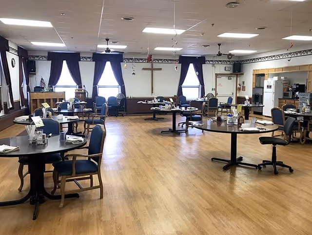 Large communal dining room with round tables and chairs on a wood floor, windows with dark curtains and a cross mounted on the far wall.