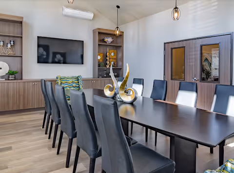 A modern conference or dining room with a long dark wooden table surrounded by black leather chairs. The room features wooden cabinetry with decorative items, a wall-mounted flat screen TV, pendant lights, and a split air conditioning unit. There are two glass-paneled doors on one side of the room.