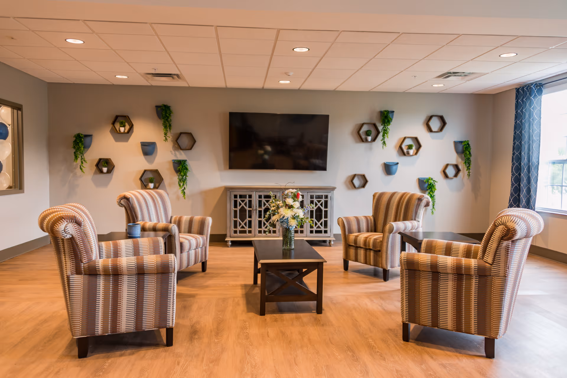 A cozy senior living common area with four striped armchairs arranged around two dark wooden coffee tables. A flat-screen TV is mounted on the wall above a decorative cabinet. The wall is adorned with hexagonal shelves and small green plants. Large windows with patterned curtains allow natural light to fill the room.