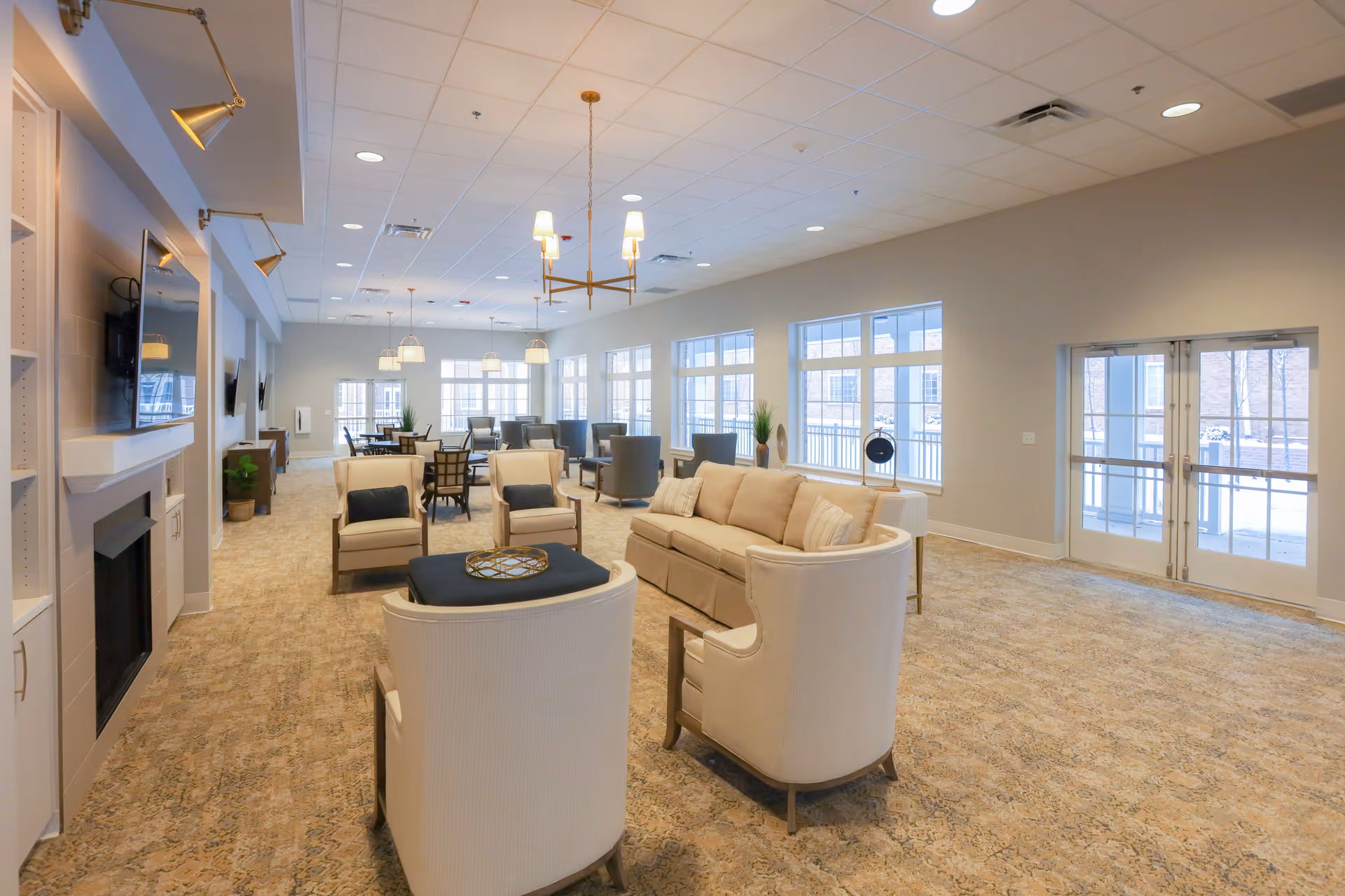 A spacious and well-lit senior living facility common area with beige carpet, multiple seating arrangements including armchairs and sofas, several dining tables with chairs, large windows letting in natural light, and modern light fixtures hanging from the ceiling. There is a fireplace with a mounted TV on the left wall and double glass doors on the right side leading outside.