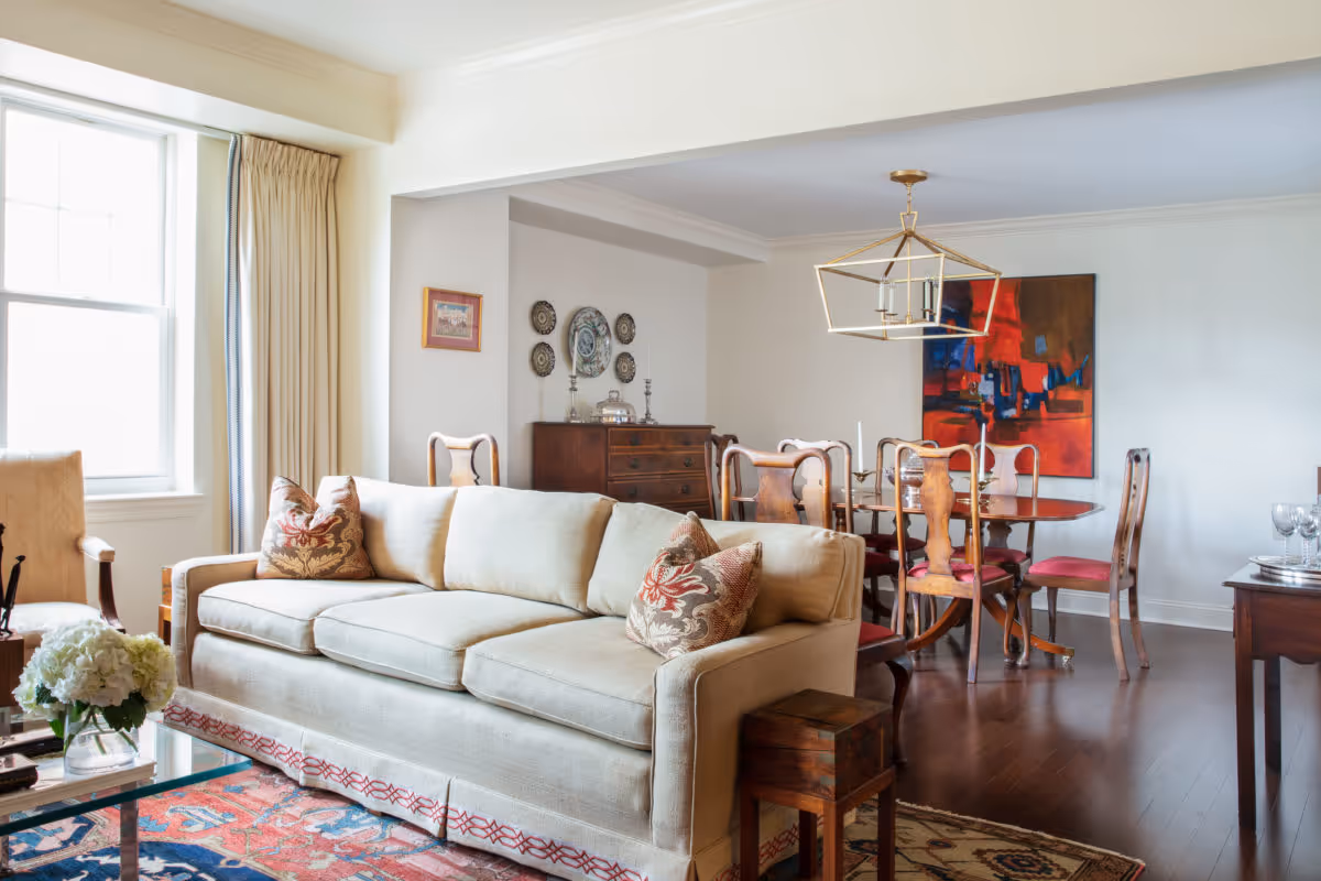 A bright living and dining area featuring a beige sofa with patterned cushions, a wooden dining table with six chairs, a modern geometric chandelier, and decorative wall art. The room has large windows with cream-colored curtains and hardwood floors with area rugs.