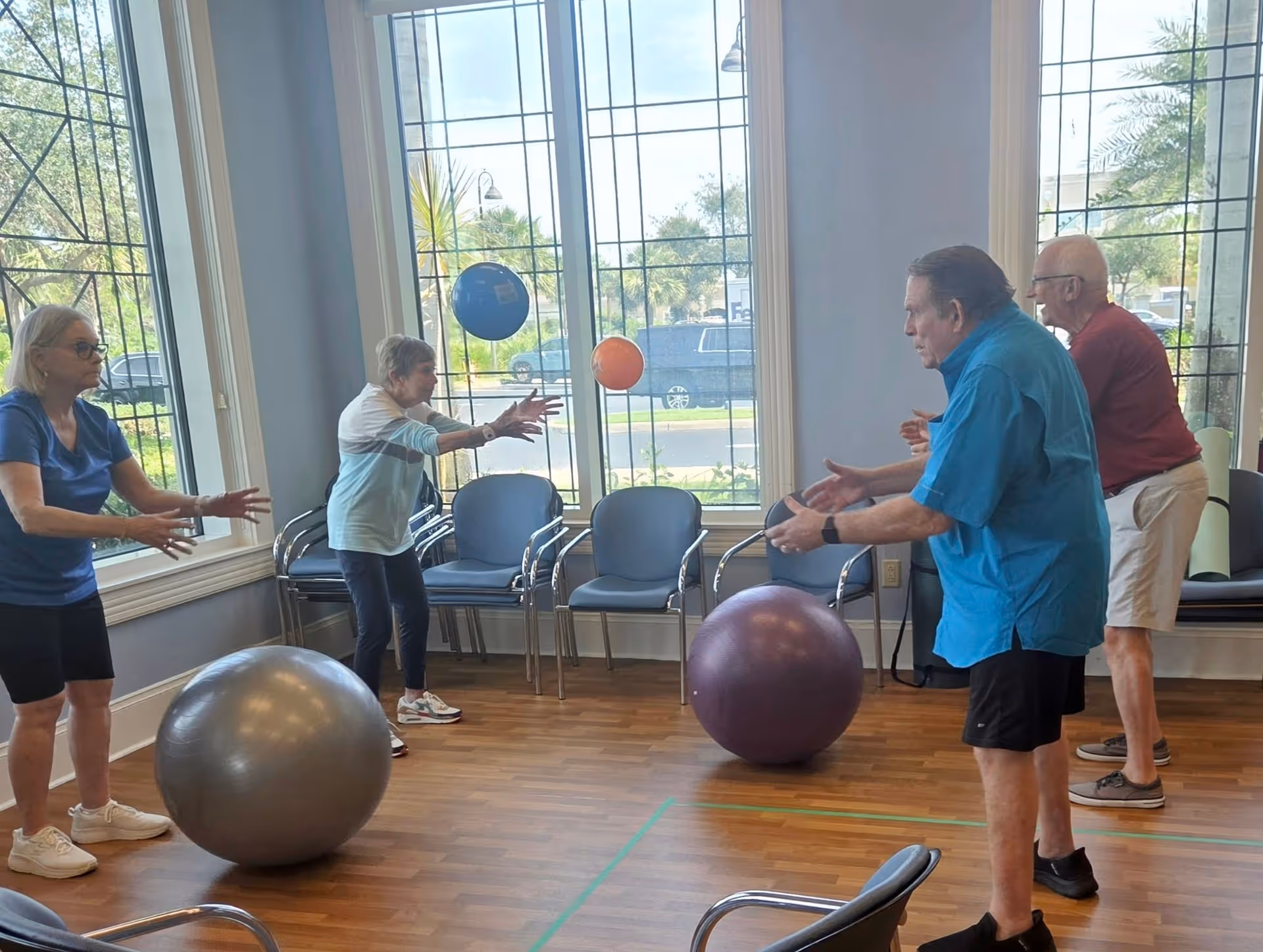 Four elderly individuals participating in a seated exercise activity indoors, tossing small colorful balls to each other in a room with large windows and several empty chairs along the walls.