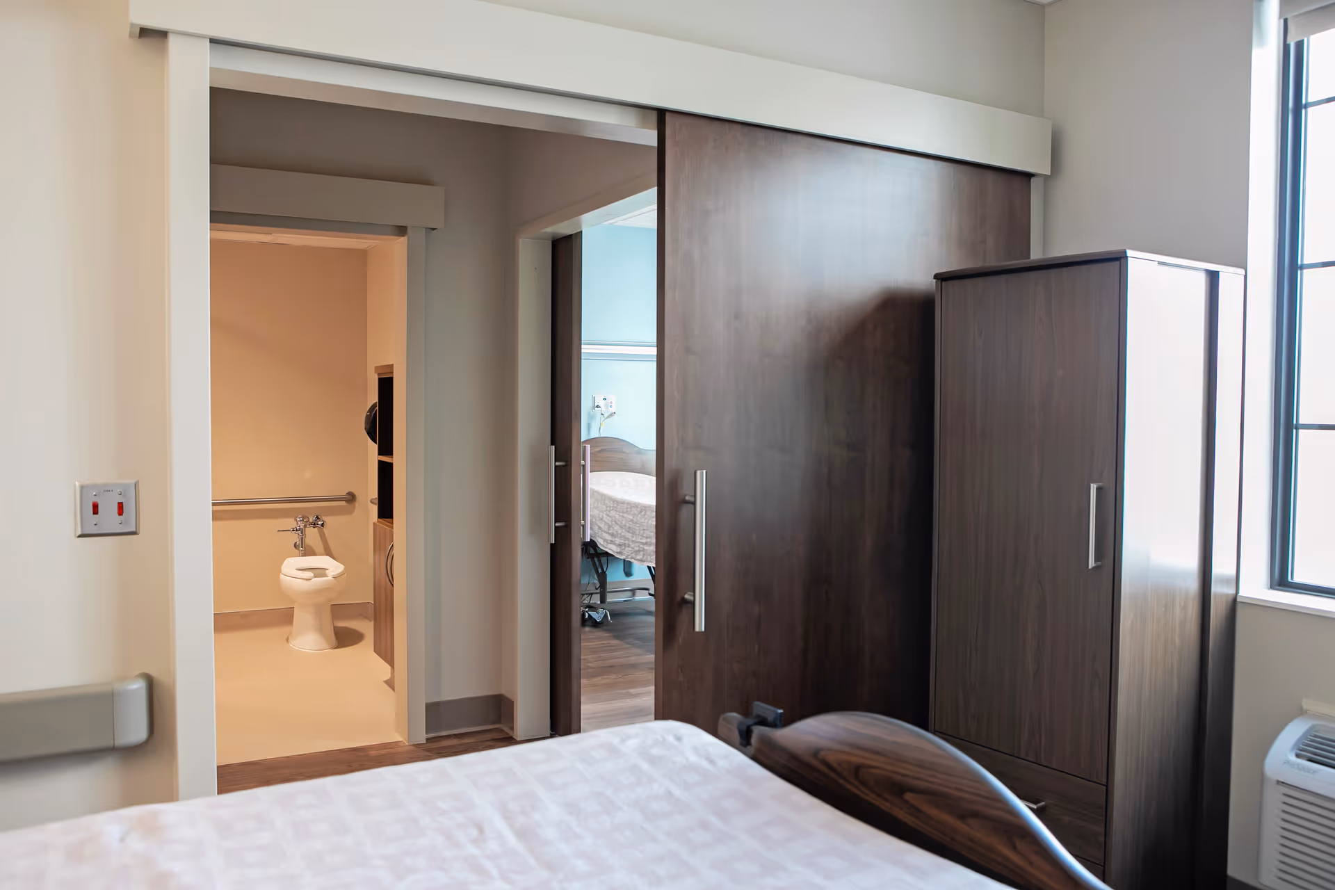 View from a bedroom in a senior living facility showing a bed with a patterned bedspread, a dark wooden wardrobe, and a sliding wooden door leading to another room with a hospital bed. To the left, there is an open doorway revealing a bathroom with a toilet and grab bars.