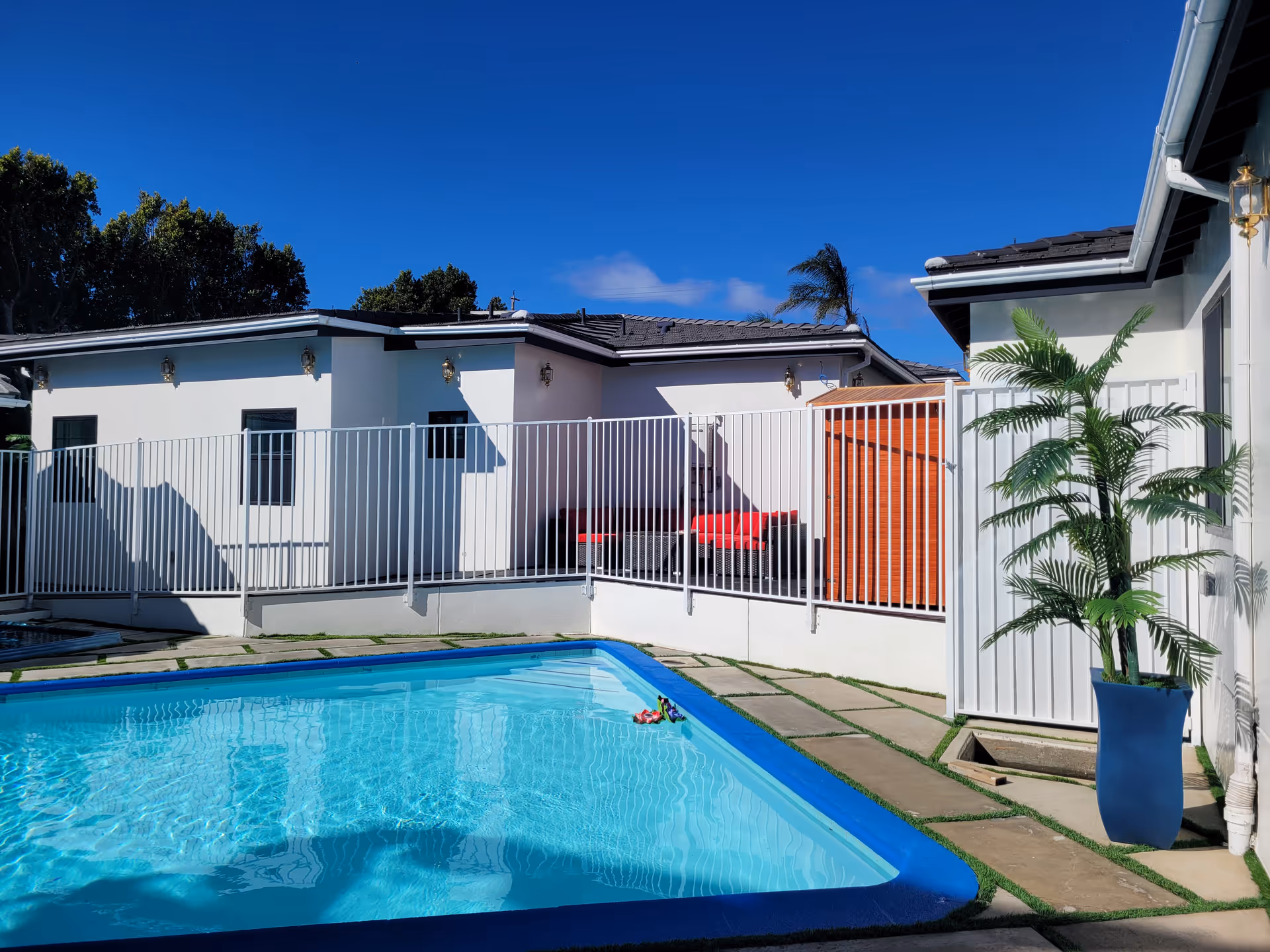 Outdoor swimming pool with clear blue water surrounded by a paved area and a white safety fence. Behind the fence, there is a white building with a dark roof, outdoor seating with red cushions, and a tall potted plant near the gate. The sky is clear and blue.